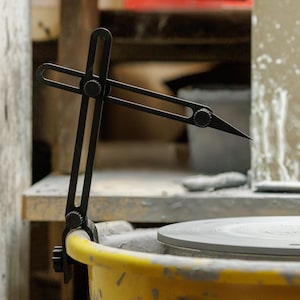 May include: A black pottery compass tool is attached to a yellow bucket, with a gray pottery wheel in the background. The tool has adjustable arms and a sharp point, used for measuring and marking clay. The setting appears to be a pottery studio.