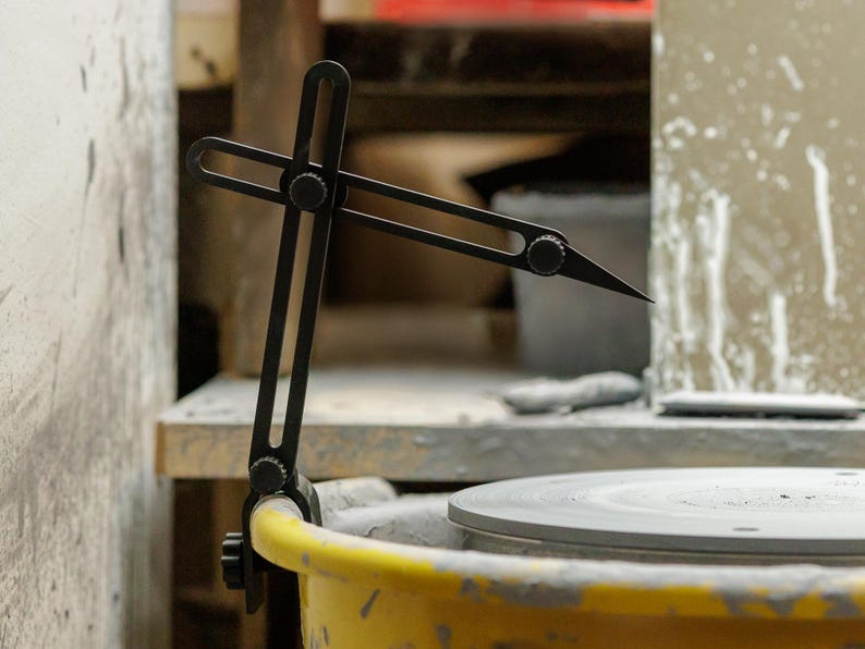 May include: A black pottery compass tool is attached to a yellow bucket, with a gray pottery wheel in the background. The tool has adjustable arms and a sharp point, used for measuring and marking clay. The setting appears to be a pottery studio.