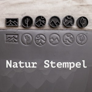 Può includere: Un set di nove timbri in gomma nera con disegni a tema natura. I timbri sono disposti su due file, con la scritta "Natur Stempel" stampata sotto. I disegni includono una catena montuosa, un cactus, un sole, una palma e una foglia.