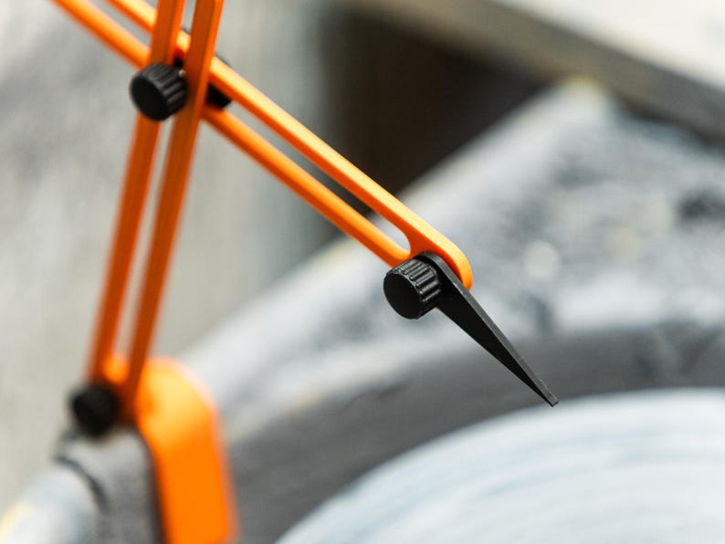 May include: Close-up of an orange and black compass tool. The tool has a bright orange frame with black adjustment knobs and a black pointed tip. The tool is designed for precise measurements and drawing circles.