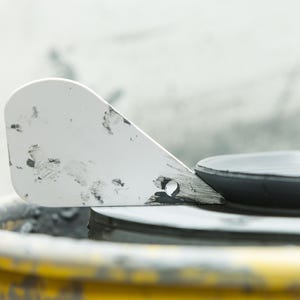 以下が含まれることがあります： 黒い汚れが付いた白い陶器のヘラが、ろくろの上に置かれています。ろくろは黄色とグレーで、その上に濃いグレーの陶器が乗っています。背景は明るいグレーです。