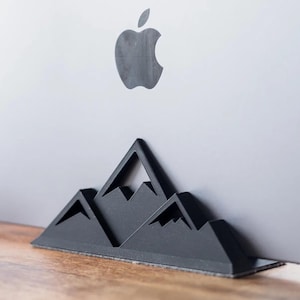 Op de afbeelding: Een zwarte, bergvormige laptopstandaard staat voor een zilveren laptop. Het Apple-logo is zichtbaar op de laptop. De standaard is gemaakt van een stevig materiaal en heeft een textuur.