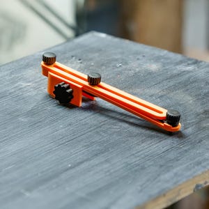 May include: An orange and black woodworking tool with adjustable knobs. The tool is resting on a dark gray surface, likely a workbench. The tool's design suggests it is used for measuring or marking.