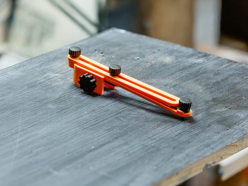 May include: An orange and black woodworking tool with adjustable knobs. The tool is resting on a dark gray surface, likely a workbench. The tool's design suggests it is used for measuring or marking.