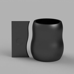 Könnte beinhalten: Eine schwarze Keramiktasse mit einer abgerundeten, organischen Form. Die Tasse steht neben einem schwarzen rechteckigen Objekt.
