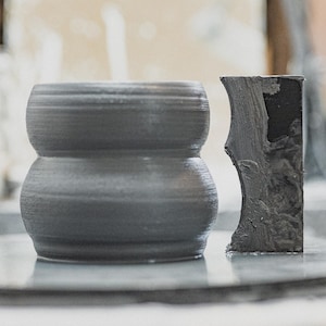 Op de afbeelding: Een grijze keramische vaas met een afgeronde zandlopervorm en een textuur. Daarnaast een donkergrijze, rechthoekige sculptuur met een ruwe, organische textuur. Beide objecten staan op een reflecterend oppervlak.