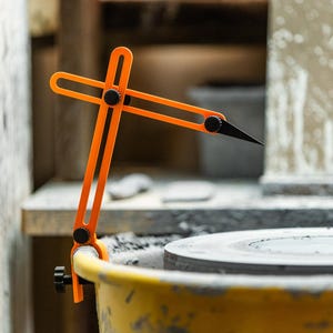 Puede incluir: Un calibrador de cerámica naranja con punta negra, montado en un cubo amarillo. La herramienta está diseñada para medir el diámetro de las piezas de cerámica. El fondo incluye un torno de alfarero y un área de trabajo.