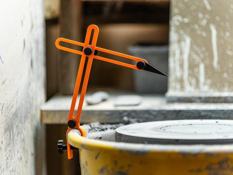 May include: An orange pottery caliper tool with a black tip, mounted on a yellow bucket. The tool is designed to measure the diameter of pottery pieces. The background includes a pottery wheel and a work area.