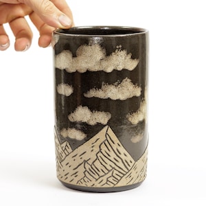 Könnte beinhalten: Ein zylindrischer Keramikbecher mit dunkelbraunem Hintergrund und Landschaftsdesign. Das Design zeigt beige Wolken und Berge. Der Becher ist etwa 15 cm hoch.