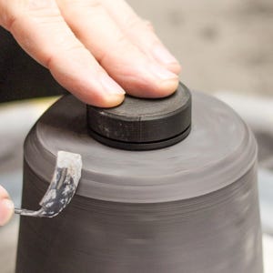Pode incluir: Um close-up das mãos de um artesão trabalhando em um objeto cilíndrico cinza escuro. Um disco preto está em cima, e uma ferramenta é usada para moldar a forma giratória. A imagem mostra a criação de cerâmica.
