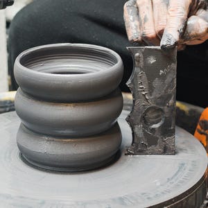 Puede incluir: Una pieza de cerámica gris sobre un torno de alfarero, con tres secciones apiladas y redondeadas. Una mano cubierta de arcilla sostiene una herramienta oscura y texturizada. La rueda y la pieza de cerámica son grises, con un fondo borroso.