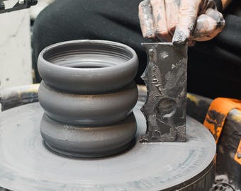 May include: A grey ceramic piece on a pottery wheel, featuring three stacked, rounded sections. A hand covered in clay holds a dark, textured tool. The wheel and the ceramic piece are grey, with a blurred background.