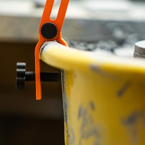 May include: Close-up of an orange and black clamp tool attached to a yellow object. The clamp has a screw for adjusting the grip. The tool appears to be used for holding or securing items during a project.