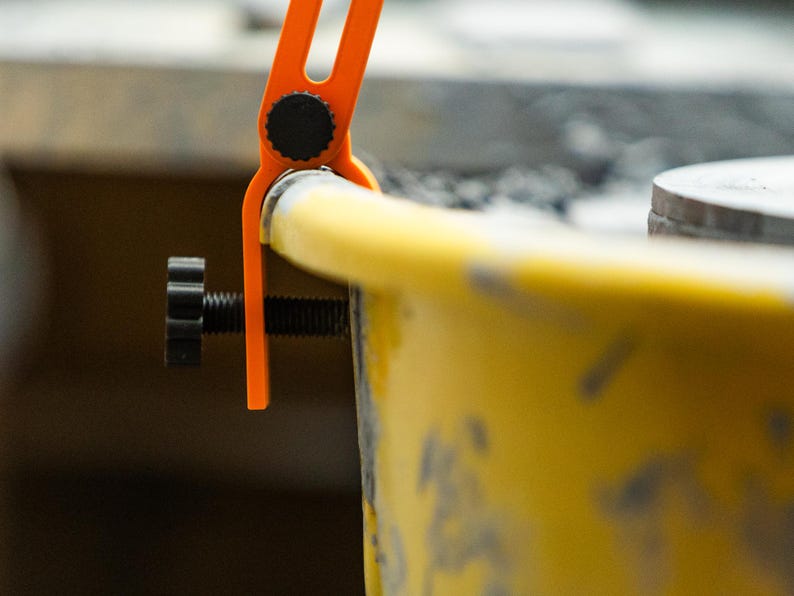 May include: Close-up of an orange and black clamp tool attached to a yellow object. The clamp has a screw for adjusting the grip. The tool appears to be used for holding or securing items during a project.