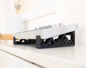 May include: A white electronic device with grey and white buttons sits on a black, mountain-shaped stand. The stand has a geometric design. The setup is on a white surface, suggesting a modern aesthetic.