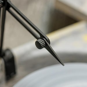 May include: Close-up of a black metal tool with adjustable arms and a pointed tip. The tool is positioned over a light gray surface, with a blurred background. The tool appears to be used for precision measurement or marking.