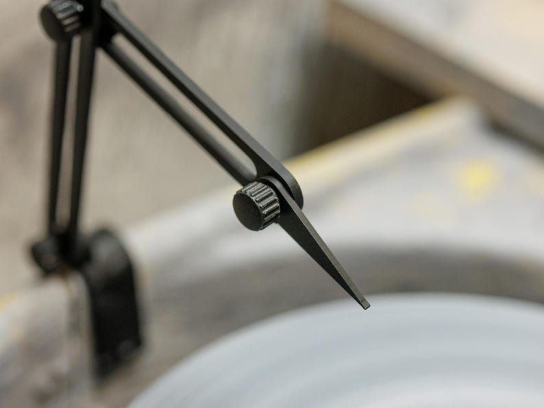 May include: Close-up of a black metal tool with adjustable arms and a pointed tip. The tool is positioned over a light gray surface, with a blurred background. The tool appears to be used for precision measurement or marking.