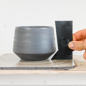 Può includere: Un vaso in ceramica grigia con una base nera si trova su un tornio da vasaio. Uno strumento in plastica nera con un foro circolare viene utilizzato per misurare la forma del vaso.