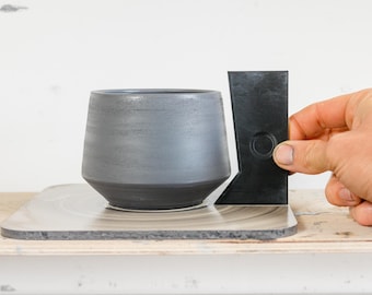 May include: A gray ceramic pot with a black base sits on a pottery wheel. A black plastic tool with a circular hole is used to measure the pot's shape.
