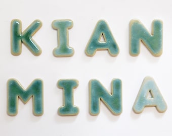 May include: A set of eight individual ceramic letters spelling KIAN MINA. Each letter features a glossy, variegated glaze in shades of teal, blue, and green with a smooth, rounded finish and a light-coloured border along the edges of the characters.