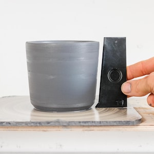 Puede incluir: Una maceta de cer&aacute;mica gris se encuentra sobre una superficie circular gris y blanca. Una herramienta de pl&aacute;stico negro con un agujero en el centro se apoya contra la maceta.