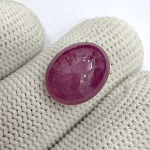 May include: A polished, oval-shaped ruby gemstone. The gem is a deep, saturated red-pink color with a smooth, reflective surface. The ruby is set against a textured, off-white background.