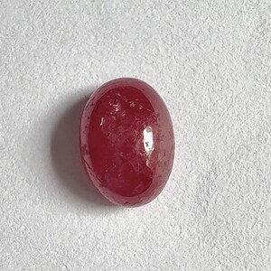 Könnte beinhalten: Ein polierter, ovalförmiger Rubin-Edelstein. Der Edelstein hat eine tiefe, satte rote Farbe mit einer glatten, reflektierenden Oberfläche. Der Stein ist vor einem weißen Hintergrund platziert, der seine Farbe und Form hervorhebt.