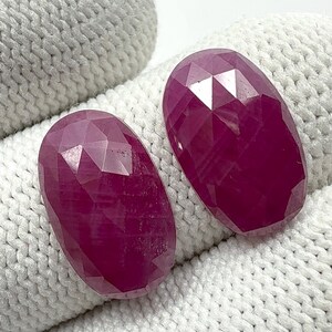 May include: Two faceted, oval-shaped rubies. The gemstones are a deep, rich red color and have a high degree of clarity. The rubies are displayed on a white, textured surface. The facets catch the light, creating a sparkling effect.