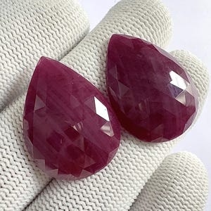 Può includere: Due gemme sfaccettate a forma di goccia, di un colore rosso intenso. Le gemme hanno una superficie lucida e riflettente, che mostra un complesso motivo di sfaccettature tagliate. Le pietre sono esposte su uno sfondo bianco strutturato.