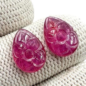 Pode incluir: Duas pedras preciosas em forma de gota de cor rosa profundo, com entalhes florais intrincados nas suas superfícies. Cada pedra apresenta um acabamento polido e translúcido com gravações detalhadas de pétalas que criam uma aparência texturizada.
