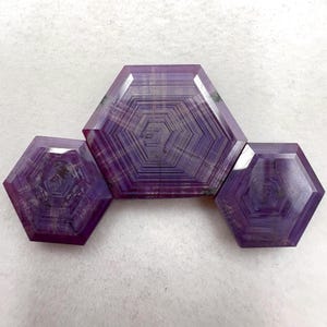 Può includere: Tre pietre preziose esagonali sfaccettate in tonalità viola e porpora intenso. Ogni pietra mostra un distinto motivo geometrico di linee esagonali concentriche che si irradiano dal centro, con superfici levigate e bordi ben definiti.