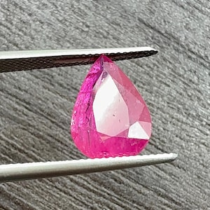 Puede incluir: Una gema rosa brillante en forma de lágrima facetada, sujeta con pinzas. La gema tiene múltiples facetas que reflejan la luz, creando un efecto brillante. La gema es de un tono rosa vibrante.
