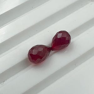 May include: Two deep red, teardrop-shaped gemstones with a faceted surface. Each stone features a polished, multi-faceted cut that reflects light, creating a sparkling appearance. The stones are positioned end-to-end in a diagonal orientation.