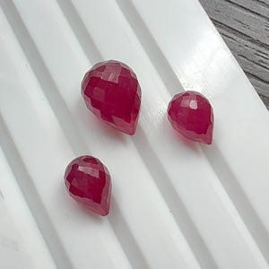 May include: Three faceted, teardrop-shaped gemstones in varying sizes. The gems are a deep, rich red color and have a polished, reflective surface. They are arranged on a white surface with parallel grooves, creating a clean and organized presentation.