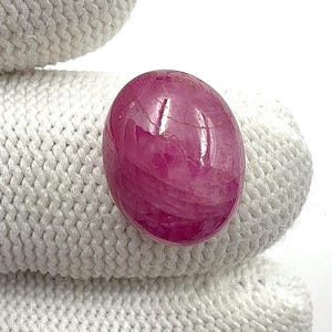 Könnte beinhalten: Ein polierter, ovaler Rubin-Edelstein. Der Edelstein hat eine tiefe, lebendige, rötlich-rosa Farbe mit einer glänzenden Oberfläche. Der Stein liegt vor einem weißen, strukturierten Hintergrund.