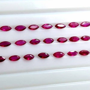 Puede incluir: Filas de gemas rojas de corte ovalado dispuestas sobre una superficie blanca. Las gemas están espaciadas uniformemente, mostrando sus facetas pulidas y su color vibrante. Probablemente rubíes, ideales para joyería.