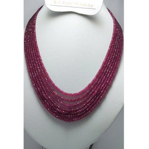 May include: A multi-strand necklace composed of small, faceted, deep red gemstones. The necklace is displayed on a white bust, with the text "B.S. RUBY TRADERS" visible on a tag at the top. The gemstones have a high luster.