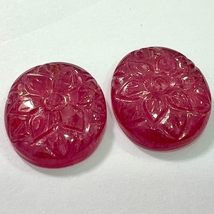 May include: Two oval-shaped, polished ruby gemstones. Each gemstone features a carved floral design. The rubies are a deep, rich red colour and appear to be translucent. The gemstones are displayed on a white surface.