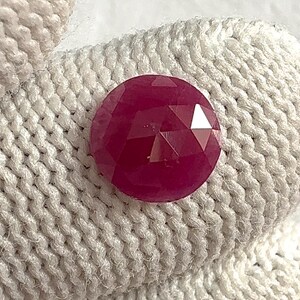 Puede incluir: Una gema facetada, redonda, de color rojo intenso. La gema tiene una superficie pulida y reflectante y está sobre un fondo texturizado blanquecino. El corte de la gema crea múltiples facetas.