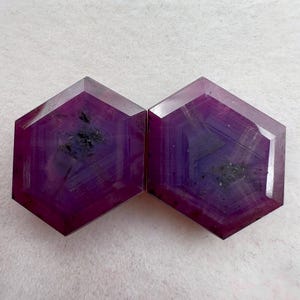 May include: Two hexagonal cut gemstones with a deep magenta and purple hue. Each stone features a faceted surface with visible internal inclusions and distinct geometric zoning patterns that radiate from the center toward the six edges.