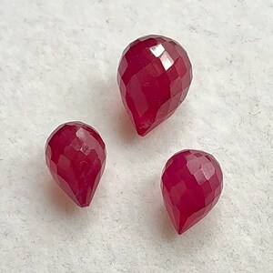 May include: Three faceted, teardrop-shaped ruby gemstones. The gemstones are a deep red color and have a polished, reflective surface. The facets create a play of light and shadow.