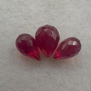 May include: Three faceted, teardrop-shaped gemstones in a deep red hue. The gems are arranged on a light gray surface, showcasing their cut and clarity. These gemstones could be used for jewelry making.