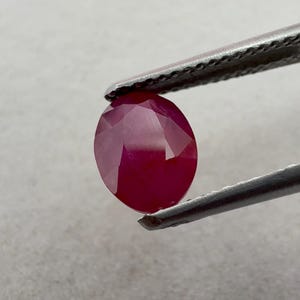 Peut inclure: Une pierre précieuse ovale rouge profond à facettes, maintenue par une pince métallique. La pierre présente une teinte cramoisie riche et saturée avec des reflets lumineux internes visibles sur sa structure polie à facettes multiples.
