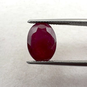 May include: An oval-cut, deep red ruby gemstone held by metal tweezers. The stone features a faceted surface that reflects light, showcasing a rich, saturated crimson colour and a symmetrical, rounded shape with clear, sharp edges.