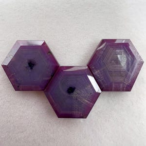 Puede incluir: Tres piedras preciosas hexagonales facetadas en un tono magenta y púrpura intenso. Cada piedra muestra un patrón geométrico distintivo de líneas hexagonales concéntricas que irradian desde un punto central oscuro.