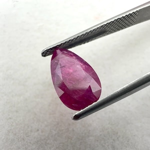 May include: A pear-shaped, faceted gemstone with a deep magenta-pink colour held by a pair of silver-toned metal tweezers. The stone features multiple triangular and kite-shaped facets that reflect light, showcasing a translucent and vibrant appearance.