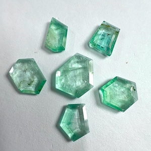 Könnte beinhalten: Sechs facettierte Smaragd-Edelsteine in verschiedenen Formen werden vor einem weißen Hintergrund gezeigt. Die Edelsteine weisen einen leuchtenden Grünton mit sichtbaren inneren Einschlüssen auf. Die Facetten fangen das Licht ein und zeigen ihre Klarheit und ihren Schliff.