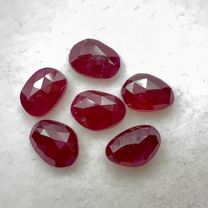 May include: Six faceted, oval-shaped, deep red gemstones are arranged on a white surface. The gems have a polished, reflective surface, and their cut reveals multiple facets. The overall arrangement suggests a collection of precious stones.