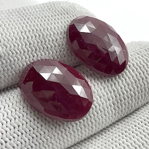 May include: Two oval-shaped, deep red gemstones with multiple facets. The gems are highly polished and rest on a light grey, textured fabric. The cut of the stones creates many reflective surfaces.