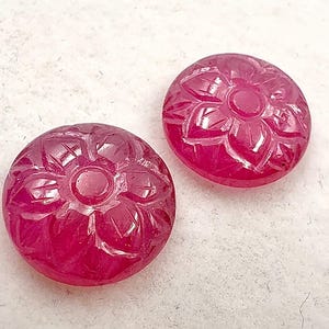 Puede incluir: Dos gemas redondas de rubí talladas. Cada gema presenta un diseño floral detallado con un elemento circular central y pétalos circundantes. Los rubíes son de un color rojo intenso y rico, y los grabados crean luces y sombras.